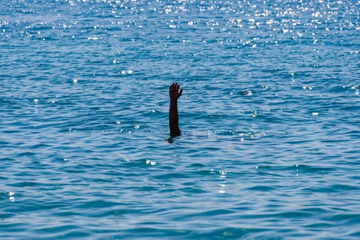 Drowning man concept, with a male raised hand with an open palm visible waving for help above a blue sea with low waves.