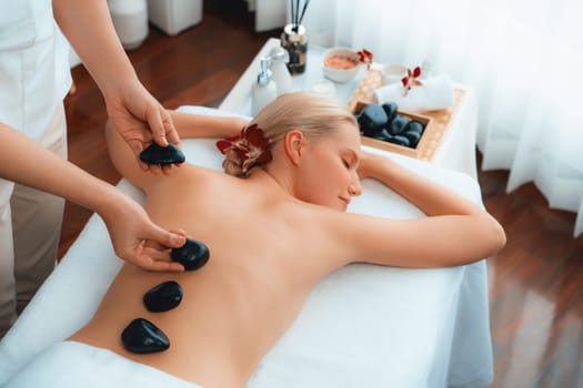 Hot stone massage at spa salon in luxury resort with day light serenity ambient, blissful woman customer enjoying spa basalt stone massage glide over body with soothing warmth. Quiescent