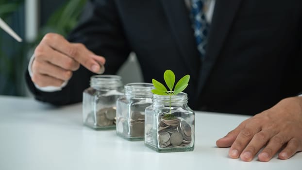 Businessman put coin to money saving glass jar at his office as sustainable money growth investment or eco-subsidize. Green corporate promot and invest in environmental awareness. Gyre