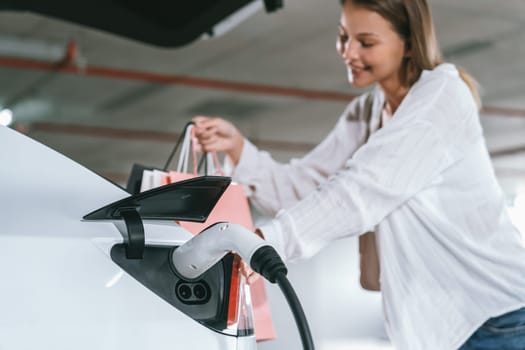 Young woman travel with EV electric car to shopping center parking lot charging in downtown city showing urban sustainability lifestyle by green clean rechargeable energy of electric vehicle innards