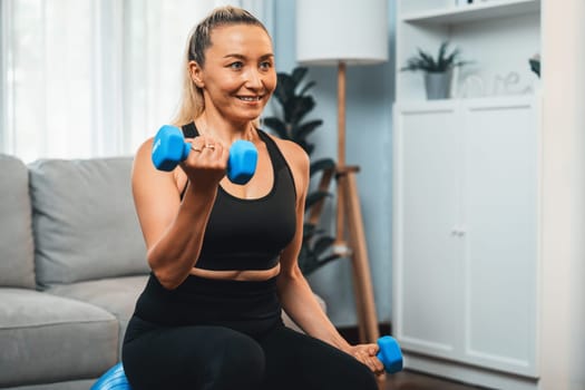 Athletic and sporty senior woman sitting on fit ball while engaging in weight lifting with dumbbell at home exercise as concept of healthy fit body lifestyle after retirement. Clout