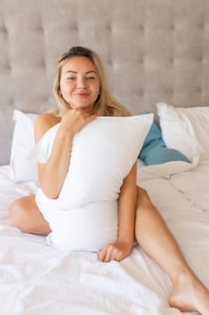 Sensual smiling young woman sitting on bed and hugging white pillow.