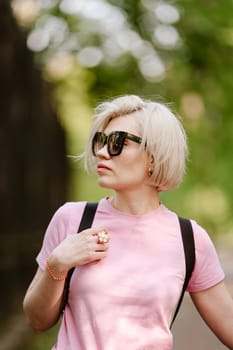 Beautiful fashionable young woman posing in the park, sunglasses, short blonde hair. Fashion summer photo. Bright colors. Nice view.