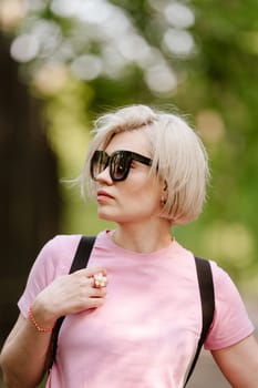 Beautiful fashionable young woman posing in the park, sunglasses, short blonde hair. Fashion summer photo. Bright colors. Nice view.