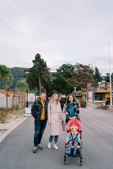 Smiling couple stands near mother with little girl in stroller on road. High quality photo