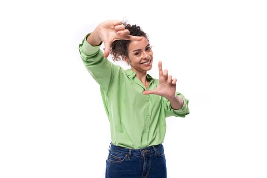 young positive brunette businesswoman with curly hair tied up in a ponytail in a light green shirt in a studio space.
