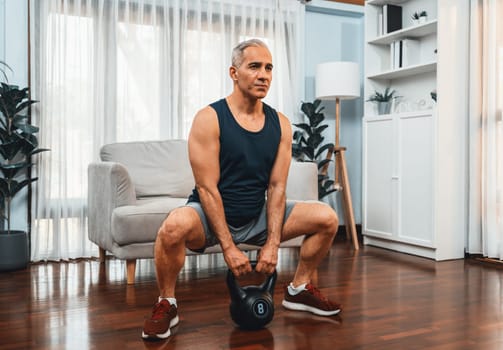 Athletic and sporty senior man engaging in leg day training session with squat and bodyweight kettle ball at home exercise as concept of healthy fit body lifestyle after retirement. Clout