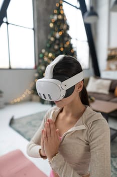 Using a virtual reality headset, a young lady in pink sportswear practices yoga near a Christmas tree. High quality photo