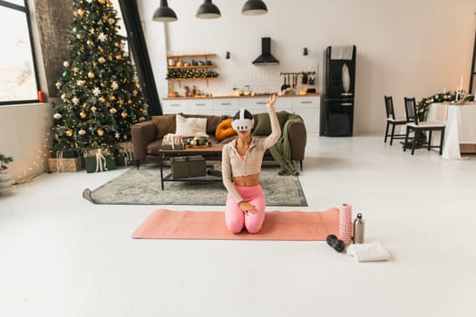A lovely young lady does her stretching routine by the Christmas tree, wearing virtual reality glasses. High quality photo
