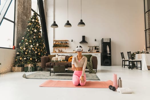 A charming young woman stretches with virtual reality glasses on in front of a Christmas tree. High quality photo