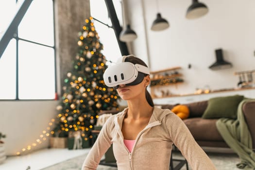 A young woman, wearing a pink sports outfit, practices yoga with a virtual reality headset near a Christmas tree. High quality photo