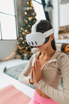 In a pink attire, a young woman practices yoga with the aid of a virtual reality headset near a Christmas tree. High quality photo