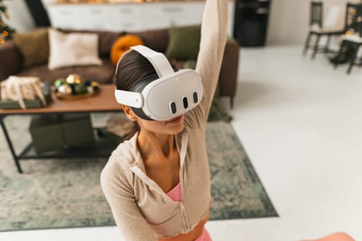 A pleasant young lady practices stretching near a Christmas tree while wearing virtual reality goggles. High quality photo