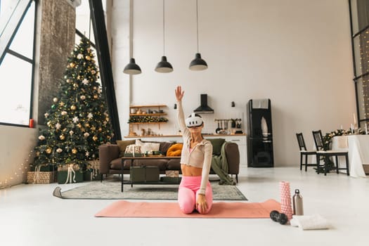 A pleasant young lady does stretching exercises beside a Christmas tree while wearing VR glasses. High quality photo