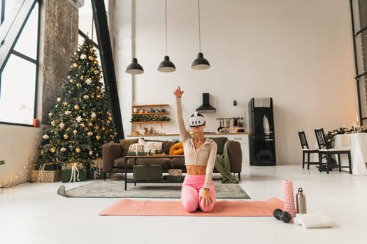 A delightful young woman engages in stretching exercises near a Christmas tree with VR goggles on. High quality photo
