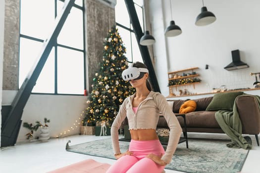 The young lady in a pink outfit practices yoga, employing a virtual reality headset against the background of a Christmas tree. High quality photo