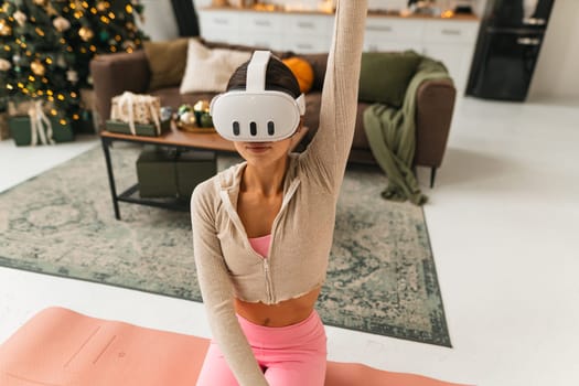 A charming young woman, wearing VR glasses, is doing stretching exercises by the Christmas tree. High quality photo