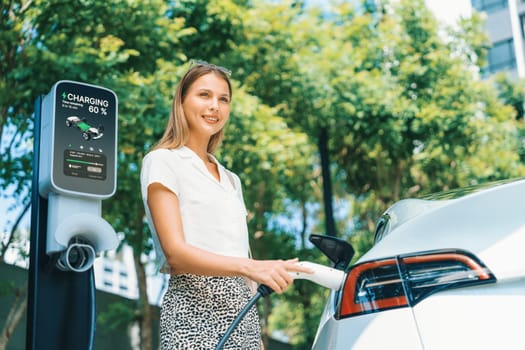 Young woman travel with EV electric car charging in green sustainable city outdoor garden in summer. Urban sustainability lifestyle by green clean rechargeable energy of electric BEV vehicle innards