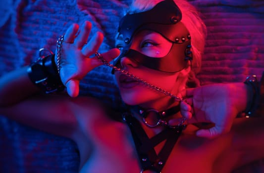 portrait of a sexy girl in a leather mask and bdsm accessories on a bed enjoying sex in orgasm with emotions on face in neon light