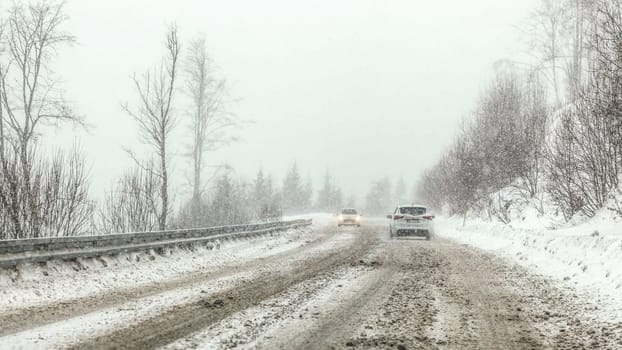 Cars driving on snow covered forest road during snowstorm, fog in disance, view from car behind. Dangerous driving conditions. 