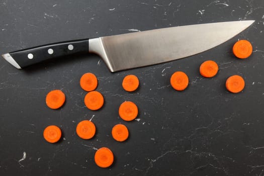 Overhead shot, carrot slices to small circles, with chef knife above, on black marble working board