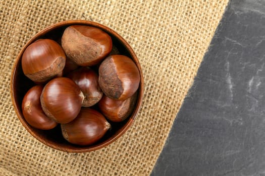 Tabletop view, small wooden bowl with chestnuts on jute cloth and slate like table