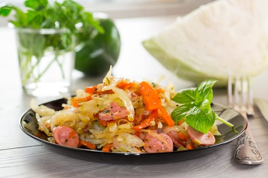 cooked fried cabbage with vegetables and sausages, in a frying pan on a wooden table.
