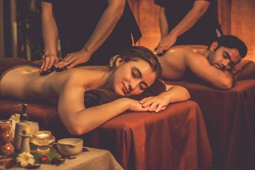 Hot stone massage at spa salon in luxury resort with warm candle light, blissful couple customer enjoying spa basalt stone massage glide over body with soothing warmth. Quiescent