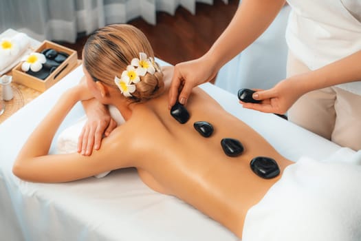 Hot stone massage at spa salon in luxury resort with day light serenity ambient, blissful woman customer enjoying spa basalt stone massage glide over body with soothing warmth. Quiescent