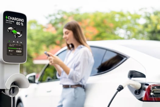 Young woman using smartphone online banking application to pay for electric car battery charging from EV charging station during vacation holiday road trip at national park or summer forest. Exalt