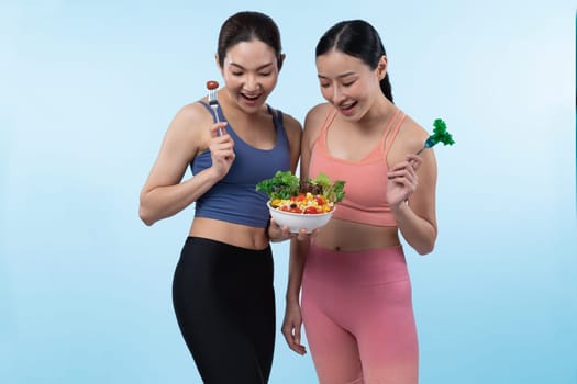 Two young sporty Asian women in sportswear holding salad bowl fill with fruit and vegetable. Natural youthful and fit body lifestyle people with balance nutrition on isolated background. Vigorous