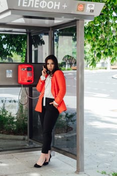 Beautiful oriental fashionable brunette woman talking in the city phone booth