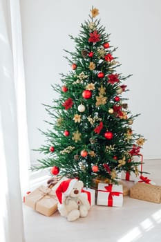 Big beautiful christmas tree decorated with beautiful shiny baubles and many different presents on wooden floor. White wall background with a lot of copy space for text. Close up.