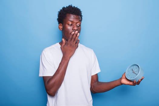 Young adult of African American ethnicity looks at the time on watch and covers his mouth in shock. Black male individual who is always on time, carrying a wall clock, and seeming anxious.