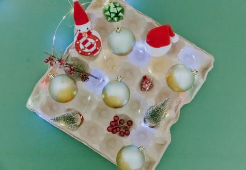 One egg carton tray with Christmas balls, burning garland, Santa hat, fir branch and Christmas decor lie in the center on a pastel green background, flat lay close-up.