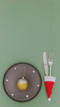 One Christmas ball with fluffy snow in a black plate with a fork and knife in a mini Santa Claus hat lie from below on a pastel green background with copy space on top, flat lay close-up.