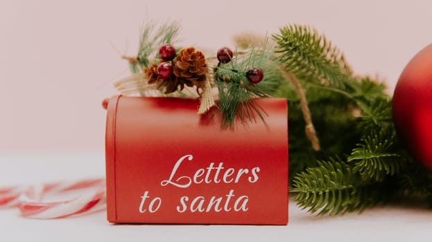 One Santa Claus mailbox with fir branches, a Christmas tree toy ball and candy lollipops stands on a light pink texture background, side view close-up.