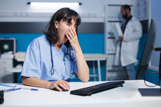 Exhausted female physician in a well-lit hospital room seated at the desk using the desktop pc. Image shows a yawning caucasian nurse working on a computer in a medical office.
