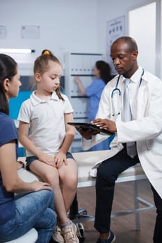 In clinic room, a doctor explains treatment to a young caucasian girl while a nurse works in background. The focus is on healthcare and medicine, with a stethoscope and digital device visible.