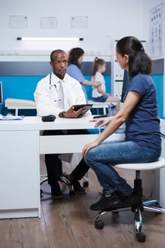 Caucasian woman in hospital room listens as African American doctor in a lab coat describes treatment procedure. Medical examination of little girl is being performed in the background by nurse.