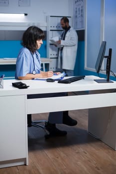 Healthcare practitioner checking patient insurance on computer and takes notes. Female physician using desktop pc in modern clinic to check and compare medical information.