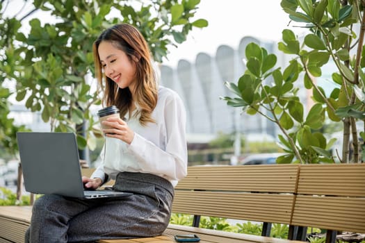 Happy Asian professional smiling businesswoman sitting alone typing computer outside street city near office, Successful business young woman working laptop outdoor corporate building exterior