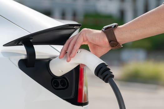 Closeup hand put EV charger to recharge electric car's battery from charging station in city commercial parking lot. Rechargeable EV car for sustainable environmental friendly urban travel. Expedient