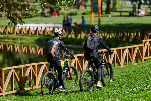 A blissful couple, adorned in professional cycling gear, enjoys a romantic bicycle ride through a park, surrounded by modern natural attractions, radiating love and happiness.