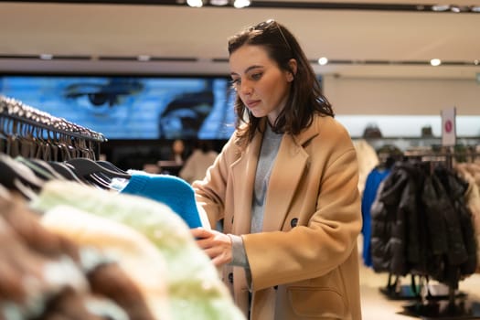 Woman picks out clothes at shopping mall dressed with thoughtful face. Female character in clothing store picks out sweater with serious face. Young adult buyer choose clothes on rack in store