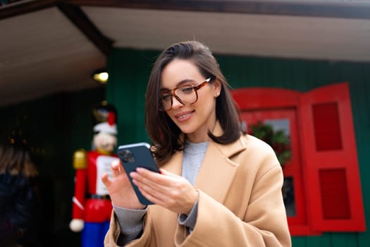 Woman on street in coat and glasses smiles while looking at phone. Happy female standing near christmas store smiling do online shopping by smartphone