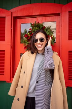 Cheerful woman smiles come out from Christmas store. Woman smiling in winter coat with sunglasses stands near Christmas store. Woman standing on street in winter coat sunglasses fixing hair. Vertical