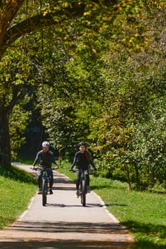 A blissful couple, adorned in professional cycling gear, enjoys a romantic bicycle ride through a park, surrounded by modern natural attractions, radiating love and happiness.