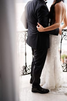 Groom hugs bride while standing on the balcony, looking at the sea. Back view. Cropped. Faceless. High quality photo