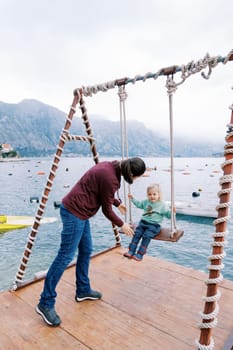 Mom swings a little girl on a swing on a pier by the sea. High quality photo